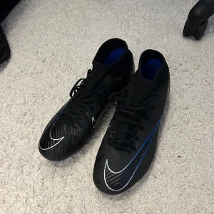 Nike Air Zoom Mercurial fotbollsskor - Snygga svarta Nike Air Zoom Mercurial fotbollsskor med blå och vita detaljer. Skorna har snörning och en strömlinjeformad design med högre skaft för extra stöd. Tillverkade i syntetmaterial för bästa prestanda på planen.