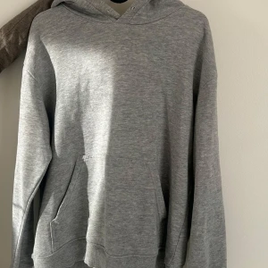 Grå hoodie med huva och ficka - En basic grå hoodie med huva och stor magficka. Tröjan har långa ärmar och ribbade muddar vid ärmslut och nederkant. Banden vid huvan är utdragna men märks knappast. Hoodien har en fin passform men kommer aldrig till användning längre.