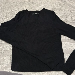 Svart ribbad tröja från Bik Bok - En svart långärmad tröja från Bik Bok i ribbad bomullskvalitet. Tröjan har en klassisk rund halsringning och sitter snyggt med en normal passform. Perfekt att styla med jeans eller kjol för en clean look.