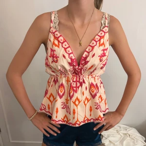Mönstrad linnetopp med korsade band - Snygg linnetopp i beige med färgstarkt rosa och orange mönster. Djup v-ringning med spetsdetaljer vid axelbanden och smala korsade axelband i ryggen. Luftig passform med resår i midjan, perfekt för varma dagar.