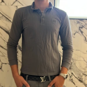 Grå långärmad pikétröja från BOSS - Snygg grå långärmad pikétröja från BOSS med klassisk krage och knappar framtill. Tröjan har diskret BOSS-logga på bröstet och är i slim fit-modell. Perfekt för dig som gillar stilrena och enkla plagg med premiumkänsla.