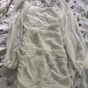 Vit kortklänning från Nelly - Supersöt vit kortklänning från Nelly med långa ärmar och snygg rynkad effekt framtill och baktill. Klänningen har en figurnära passform och är gjord i ett lätt material som känns luftigt. Perfekt för dig som vill ha en stilren och trendig look.