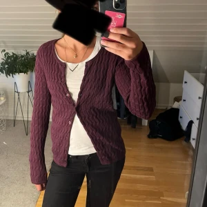 Vinröd kofta från Boomerang - Säljer en vinröd kofta från Boomerang i stickat material. Koftan har ett snyggt kabelstickat mönster, rund halsringning och knäppning framtill. Perfekt att slänga över en t-shirt för en chill och stilren look.