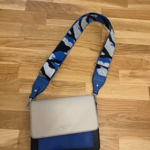 Axelväska Art Street London blå/grå - Snygg axelväska från Art Street London i skinnimitation med beige lock, blå och svarta detaljer samt bred axelrem i blått, svart och silver med camouflage-mönster. Väskan har flera fack inuti och cool kontrast mellan färgerna. Pris kan diskuteras 