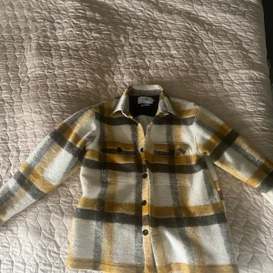 NN07 jacka - Overshirt i mjuk ullmix från NINE med rutigt mönster i vitt, grått och gult. Klassisk krage, knäppning framtill och två bröstfickor med knappar. Perfekt lager-på-lager-plagg för höst och vår. Liten i storleken, passar som en large.