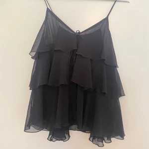 Svart volangig linnetopp från Zara - Supersöt svart linnetopp från Zara med tunna axelband och flera lager volanger. Toppen har en lätt och luftig känsla, perfekt för varma dagar. Snygg knytdetalj framtill och flowig passform som ger en cool och trendig look. Använd en gång.