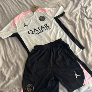 PSG x Jordan vit och svart fotbollströja - Snygg PSG x Jordan fotbollströja i vitt med svarta och rosa detaljer. Tröjan har korta ärmar, rund hals och Paris Saint-Germain-logga på bröstet. Tillverkad i lätt och ventilerande polyester, perfekt för träning eller match.