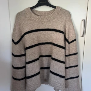 Beige stickad randig tröja  - Beige stickad tröja med svarta horisontella ränder och rund hals. Tröjan har en loose passform och långa ärmar. Använd några gånger men är nästan som ny. Perfekt till vintern eller sena sommarkvällar.