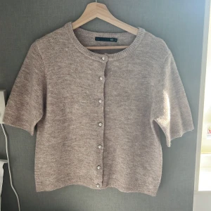 Beige stickad kofta från lager 157 - Supermjuk och mysig beige stickad kofta från 157 med kort ärm och pärlknappar framtill. Koftan har en rund halsringning och ribbade kanter för en chill och stilren look. Perfekt att slänga över en topp när det är lite kyligt ute.