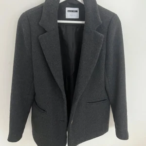 Mörkgrå kavaj från Veromoda - Snygg mörkgrå kavaj från Veromoda med klassisk krage och två knappar framtill. Jackan har två fickor och är helfodrad, perfekt för dig som vill ha en stilren och enkel look. Passar till både jeans och kostymbyxor.