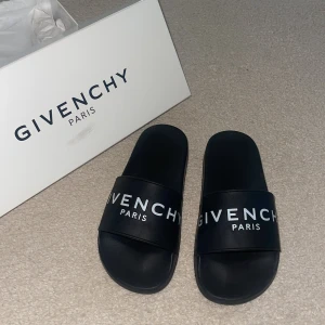 Svarta slides från Givenchy Paris - Svarta slides från Givenchy Paris med vit logga över foten. Snygg och clean design med platt sula och öppen tå. Tillverkade i syntetmaterial, perfekta för sommaren eller chill dagar. Kommer med originalkartong.