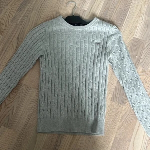 Grå kabelstickad tröja från Gant - Snygg grå kabelstickad tröja från Gant i stretchig bomull. Klassisk rund halsringning, diskret Gant-logo på bröstet och ribbade muddar. Perfekt för lager-på-lager och chill stil under höst och vinter.