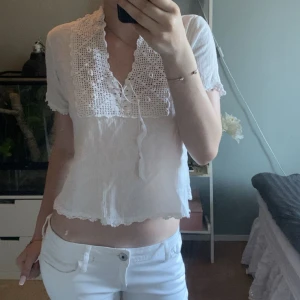 Vit blus med virkade detaljer - Supersöt vit blus med korta ärmar och croppad passform. Blusen har en stor virkad krage och virkade detaljer framtill med snörning i urringningen. Gjord i ett lätt och luftigt bomullstyg, perfekt för varma dagar. Ger en romantisk och bohemisk vibe.