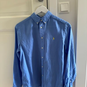 Blå skjorta från Ralph Lauren - Klassisk blå linneskjortaskjorta från Ralph Lauren med button-down krage och broderad gul logga på bröstet. Skjortan har långa ärmar, vita knappar och är tillverkad i linen. Perfekt för en clean och stilren look.