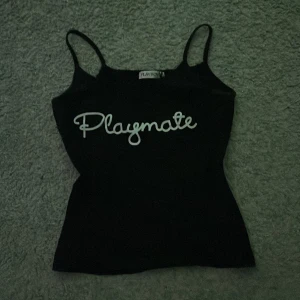 Svart Playmate linne från Playboy - Svart linne från Playboy med smala axelband och vit text 'Playmate' framtill. Sitter tight och har en klassisk, rak form. Perfekt för dig som gillar en enkel men ändå statement look.