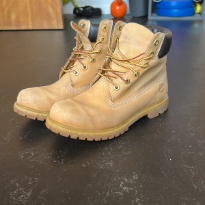 Beiga Timberland boots  - Klassiska Timberland boots i beige nubuckskinn med robust snörning och grov sula. Skorna har vadderad svart krage runt ankeln och ikoniska Timberland-loggan på sidan. Perfekta för höst och vinter.