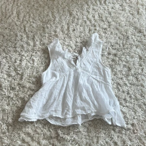 Vit blus med volang från Gina Tricot - Supersöt vit blus från Gina Tricot i luftig bomull. Blusen har volangdetaljer vid axlarna, v-ringning och knytning framtill. Perfekt för varma dagar och ger en romantisk vibe med sitt tunna, flowiga tyg.