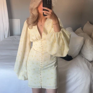 Gul Adoore klänning - Superfin ljusgul kortklänning från Adoore med små blommor, djup v-ringning och markerad midja. Klänningen har stora puffärmar och knappar framtill som ger en unik look. Perfekt för sommarens alla tillfällen! Endast använd 1 gång, storlek 36, men passar 34! Nypris 1500