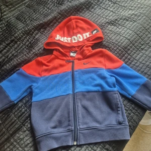 Nike hoodiejacka med färgblock - Nike hoodiejacka med dragkedja och huva. Snygg färgblock-design i rött, blått och mörkgrått. Tryckt 'JUST DO IT.' på huvan och klassisk Nike-logga på bröstet. Mjuk och skön i bomullsmix, perfekt för chill dagar eller när du är på språng.