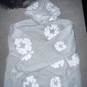 Grå hoodie från Denim Tears - Säljer en grå hoodie från Denim Tears med vita blommönster. Den har en klassisk passform med en stor ficka framtill och en bekväm huva. Perfekt för en avslappnad stil.