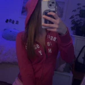 Rosa zip hoodie från Abercrombie & Fitch - Säljer en rosa hoodie från Abercrombie & Fitch med dragkedja och huva. Jackan har vita tryckdetaljer framtill och är i mjukt material, perfekt för chill dagar. Klassisk college-vibe och snygg passform.