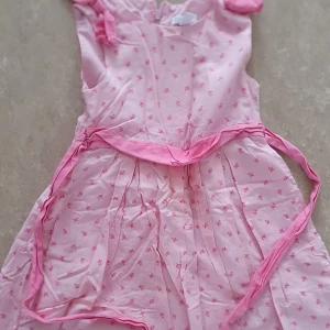 Klänning - Rosa klänning med små blommiga mönster och knytband i midjan. Klänningen har korta ärmar och är gjord i ett lätt tyg, perfekt för varma dagar. Den har en feminin och gullig stil med fina detaljer.