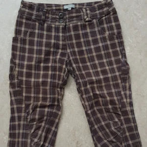 Byxor - Bruna byxor med rutigt mönster från Kids by Lindex. Byxorna har raka ben, två knappar i midjan och klassiska fickor fram. Snyggt rutmönster i brunt och beige med tunna blå linjer. Perfekta för dig som gillar en cool och avslappnad stil.