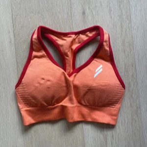 Orange sportig linnetopp med röd kant - Sportig orange linnetopp med röda kanter och vit logga framtill. Toppen har bred resår nedtill och är ärmlös med brottarrygg, perfekt för träning och aktiva dagar. Snygg och bekväm passform som håller allt på plats.