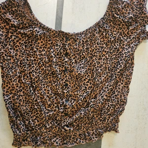 Leopardmönstrad topp från Lindex - Snygg leopardmönstrad topp från Lindex i bruna och svarta toner. Toppen har resår nedretill och knappar framtill för en trendig look. Perfekt för dig som vill sticka ut med ett djurmönster. Materialet är mjukt och skönt mot huden.
