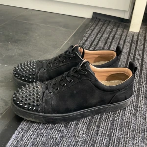 Christian Louboutin svarta sneakers med nitar - Svarta sneakers i skinn från Christian Louboutin med ikoniska röda sulor och svarta nitar på tån. Skorna har snörning, rund tå och en cool, edgy vibe. Insidan är beige och sulan har Louboutin-loggan i rött. Perfekta för dig som vill sticka ut. Storlek 39 men passar 40. De skulle också kunna funka på 37/38 men då är de ganska stora. Lite smutsiga men kan göra rent när nån köper! Tar också byten.