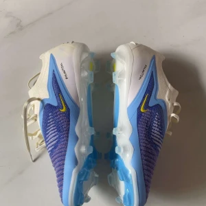 Nike Phantom fotbollsskor - Nike Phantom fotbollsskor i blått och vitt med gula detaljer. Ovandelen är stickad och har snörning för bra passform. Sulan är ljusblå med dobbar för optimalt grepp på planen. Perfekta för dig som vill sticka ut på matchen.