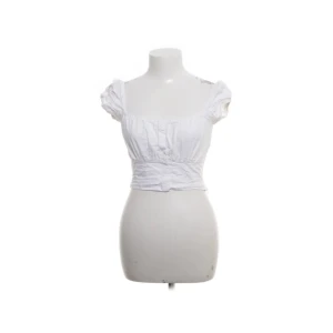 Vit croppad blus med puffärm - Supersöt vit croppad blus med korta puffärmar och smockad midja. Toppen har en feminin v-ringning och knappar framtill. Perfekt för varma dagar och ger en romantisk vibe till din outfit.