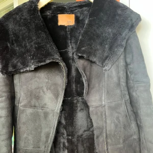 Svart pälsjacka från EMU Australia - Säljer en svart pälsjacka från EMU Australia med stor huva och mjukt foder. Jackan har markerade sömmar, dragkedja och är tillverkad i skinn med päls på insidan. Perfekt för kalla dagar när du vill vara både varm och snygg.