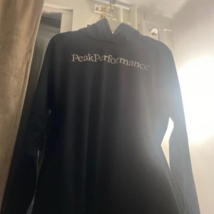 Svart hoodie från Peak Performance - Svart hoodie från Peak Performance med broderad logga framtill. Klassisk modell med huva och långa ärmar, perfekt för en chill och sportig stil. Mjuk och skön, troligtvis i bomullsmix. Snygg att matcha med jeans eller träningsbyxor.