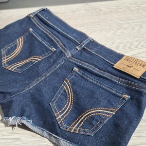 Mörkblå jeansshorts från Hollister - Snygga mörkblå jeansshorts från Hollister med klassiska bakfickor och kontrastsömmar. Modellen är lågmidjad med råa, fransiga benslut för en avslappnad vibe. Perfekta för sommardagar och festivaler. Ikonisk Hollister-logga på bakfickan och läderpatch i midjan. Köpta secondhand