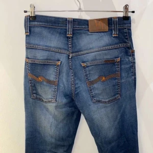 Blå jeans från Nudie Jeans - Säljer ett par klassiska blå jeans från Nudie Jeans med snygga slitningar och kontrastsömmar. Jeansen har raka ben, fem fickor och ikonisk broderad detalj på bakfickorna. Perfekta för dig som gillar en avslappnad men stilren look.