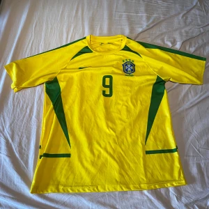 Brasilien Ronaldo 9 fotbollströja Nike - Gul Brasilien fotbollströja från Nike med gröna detaljer, nummer 9 och Ronaldo på ryggen. Klassisk design med korta ärmar, rund hals och broderat landslagsmärke på bröstet. Tillverkad i lätt och ventilerande polyester, perfekt för fotboll eller samling.