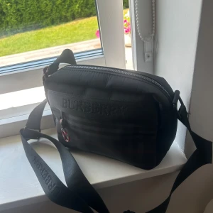 Svart axelväska från Burberry - Snygg svart axelväska från Burberry med diskret logga på framsidan och justerbar axelrem. Väskan har dragkedja upptill och är tillverkad i slitstarkt syntetmaterial. Insidan är fodrad med Burberry-mönstrat tyg och har en innerficka för småprylar.