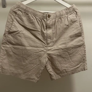 Beige linneshorts från H&M - Snygga beige shorts från H&M i regular fit med elastisk midja och knäppning fram. Tillverkade i svalt linne, perfekta för varma dagar. Två sidofickor och två bakfickor med knapp för en clean look. 