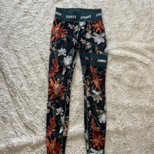 Blommiga leggings från Stronger XS - Snygga leggings från Stronger i storlek XS med färgglatt blommönster i orange, vitt och grönt. Hög midja med logotyp i midjebandet. Tillverkade i återvunnen polyester och elastan för en stretchig och bekväm passform. Perfekta för träning eller chill.