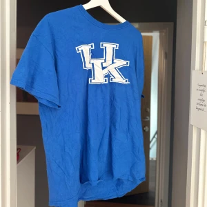 Blå Kentucky Wildcats t-shirt - Blå t-shirt med stort vitt UK-tryck på bröstet från Kentucky Wildcats. Klassisk passform med rund hals och korta ärmar. Perfekt för dig som gillar sportig och avslappnad stil.