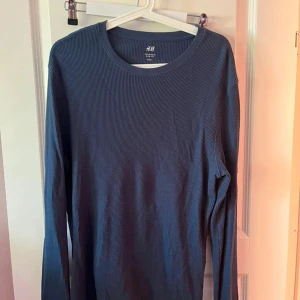 Blå ribbad långärmad tröja H&M - En stilren blå långärmad tröja från H&M med ribbad struktur. Tröjan har rund halsringning och är tillverkad i mjuk bomull, perfekt för lager-på-lager eller chill dagar. Enkel att matcha med jeans eller joggers.
