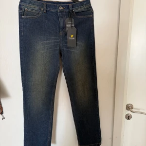 Mörkblå jeans från Lyle & Scott - Snygga mörkblå jeans från Lyle & Scott med klassisk femficksdesign och gul logga på bakfickan. Jeansen har en rak passform och subtila slitningar framtill och vid knäna. Perfekta för dig som gillar stilrena och tidlösa jeans.