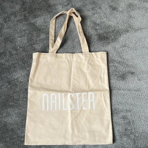 Beige shoppingväska från Nailster - Enkel och stilren shoppingväska i beige bomull med vita tryckta bokstäver 'NAILSTER' på framsidan. Väskan har långa handtag som gör den smidig att bära på axeln. Perfekt för att ta med till skolan, stan eller gymmet.
