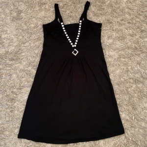 Svart kort klänning med silvriga detaljer - Svart kortklänning från Kozz Style med breda axelband och djup v-ringning dekorerad med silvriga nitar och en fyrkantig detalj. Mjuk och följsam passform, perfekt för dig som gillar en stilren look med lite edge.