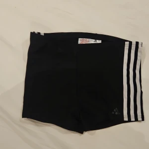 Svarta Adidas hotpants med vita ränder - Svarta sportiga hotpants från Adidas med klassiska tre vita ränder längs sidorna och Adidas-logga framtill. Tillverkade i ett lätt och stretchigt syntetmaterial som passar perfekt för träning eller sport. Snygg och enkel design med normal passform. Angiven stl är i barn och motsvarar stl 164 alternativ xxs.