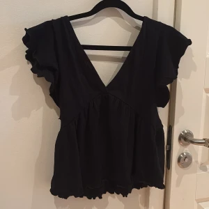 Svart blus med volangärm och v-ringning - Supersöt svart blus med djup v-ringning både fram och bak, volangärmar och knyt i ryggen. Blusen har en luftig och flowy passform med lätt rynk vid midjan och vågig kant nedtill. Perfekt för dig som gillar en romantisk och trendig stil.