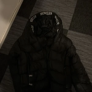 Svart Moncler dunjacka med huva - Säljer en svart dunjacka från Moncler med stor huva och tydlig logga på ärmen. Jackan har dragkedja framtill och coola detaljer med Moncler-tryck på huvkanten. Perfekt för kalla dagar när du vill hålla stilen.