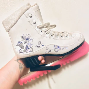 Vita konståkningsskridskor med blommor - Vita konståkningsskridskor från Nancy i storlek 35, dekorerade med handritade blommor i blått och grått på sidan. Skorna har snörning, mjuk päls vid skaftet och rosa skydd på skenan. Perfekta för dig som vill sticka ut på isen!