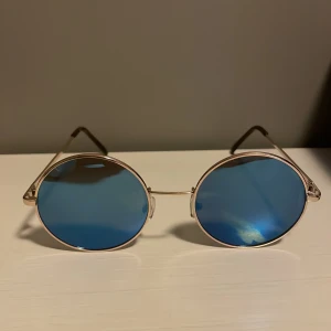 Runda solglasögon med blå spegelglas - Snygga runda solglasögon med tunna metallbågar i silver och blå spegelglas. Klassisk retrostil med justerbara näskuddar och smala skalmar. Perfekta för dig som vill sticka ut med en cool och tidlös look.